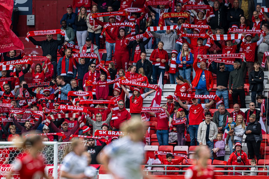 Brann-fans støtter sine spillere før et møte med Rosenborg i Toppserien 2024, på Brann Stadion. Står med skjerf og synger.